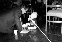 Dr. Maurer on hands and knees is looking at a baby sitting on the floor, chewing on the top of his cane.