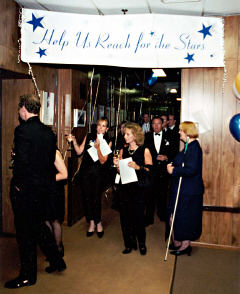 We can see people walikng toward the camera, programs in hand.   A blue and white signwith stars and the words "Help Us Reach for the Stars,"hand across the hallway.