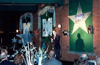 A m,an stands speaking at the podium against the east wall of the skydeck.   Behind him is a large silver star containing a picture of the model of the National Research and Training Institute for the Blind.   Star-shaped helium balloons flank the star.    In the foreground one of the star tables can be clearly seen.