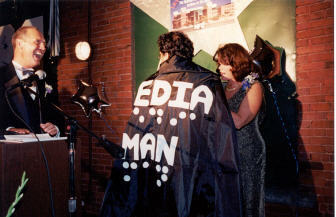 A man stands with his back largely to the camera.   Betsy Zaborowski has just put a cape on him and is tying it in front of him.   Much of the print and braille can be seen on his back,