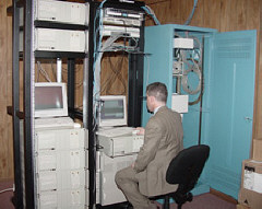 On March 1, 2002, NFB-NEWSLINE went nationwide.   Picyured here is John Berggren, NFB-NEWSLINE Systems Administrator seated in front of racks holding the ten NFB -NEWSLINE computers.  On the right is a cabinet containing the interface between incoming fiber optic cable and a DS-3 line.   The cednter rack houses the multiplexer that splits the DS-3 into the T-1 lines.   Initially these lines will ptovide simultaneous service for up to 322 newspapers readers.