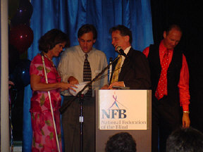 Left to right are Betsy Zaborowski, Erik Weihenmayer, and President Marc Maurer.  Jim Gashel looks on as Erik pulls a name from an NFB plastic hard hat.   The drawing winner was to receive a $1,000 bill.