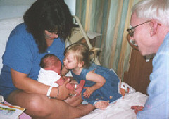 Caroline leans over to kiss baby Meghan sitting i n Alice's lap while proud daddy Michael looks on.