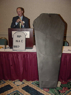 Jim Gashel addresses the Friday evening meeting.  In front of him is the NAC headstone propped in front of the podium, and the NAC casket leaning against the table.