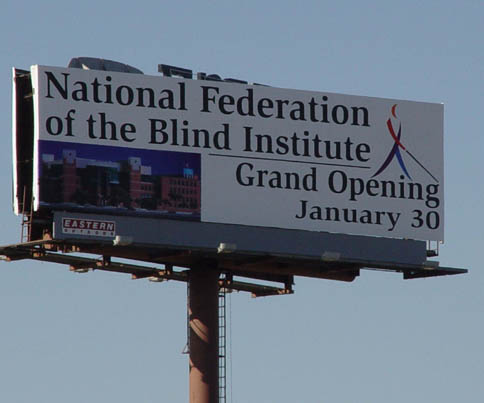 On the billboard pictured here the words "National Federation of the Blind" appear on the left with a full-color Whozit on the right.   Below the words is a picture of the model of the Institute and at the bottom right are the words, "Grand Openng January 30th"