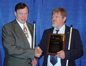 Steve Benson and Jerry Whittle shake hands.   Jerry Whittle is holding his Blind Educator of the Year plaque.