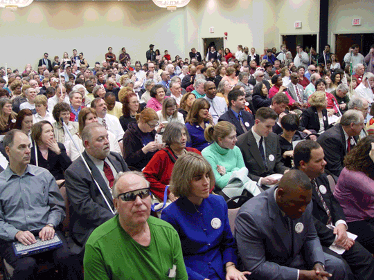 The photograph shows a large meeting room filled with people. 