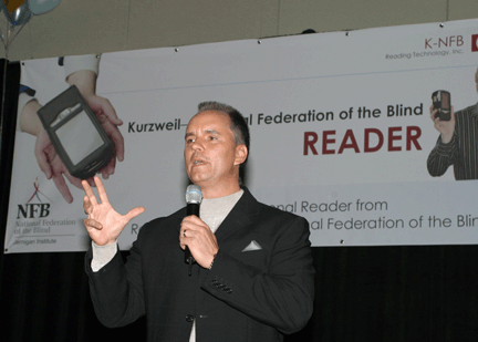 Master of Ceremonies Gerry Sandusky stands on the stage in front of the Kurzweil-National Federation of the Blind Reader banner.