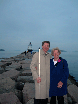 John and Cindy Par&eacute; stand on a jetty constructed of boulders. A lighthouse is visible behind them.
