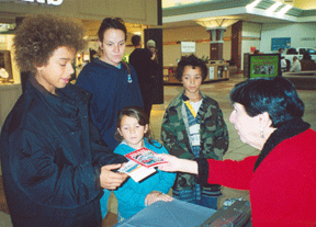 Delores Reisinger, member of the Des Moines Chapter of the NFB of Iowa, distributes Kernel Books and Braille alphabet cards at the Lindale Mall in Cedar Rapids to a mother and three children who stopped by her literature table to learn more about the NFB.