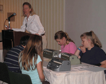 Michael Taboada, Hannah Weatherd, Megan Bening, and Kyra Sweeney at table during team portion of MathCounts, while Kristin Chandler of MathCounts gives instructions