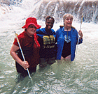 Two senior blind women stand in a waterfall with their friend.