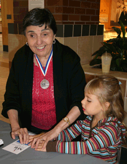 Proudly wearing her NFB March for Independence medallion, Dolores Reisinger demonstrates the Braille code to six-year-old Alexa Martin using a Braille alphabet card during one of her many MTBM outings. This one was at Lindale Mall in Cedar Rapids, Iowa.