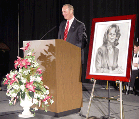 Standing at the podium in Members Hall, Jim Gashel pays loving tribute to his late wife. A beautiful floral arrangement is massed in front of the podium.