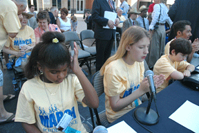 March&eacute; Daughtry, Hannah Weatherd, Jordan Richardson, and Vejas Vasiliauskas are seated at the head table on the rally stage. They presented a resolution on the importance of Braille and the NFB as tools to help their generation gain a quality education and solid employment. Hannah Weatherd is speaking into the microphone while the others listen.