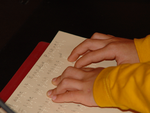 The little hands of Brandon Pickrel, a child who attended the NFB of Maryland convention in 2007, are pictured here reading Braille. The print text of what he is reading can be seen above the Braille.