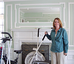 Lori Brown stands in Mrs. Pillsbury�s bedroom, which is currently filled with exercise equipment. Behind her are the painted mantle and the marble fireplace. Also visible are the beautifully painted wainscoted and paneled walls of the room.