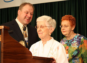 Ruth Bogia stands at the podium with Nancy Amick behind her and Gary Wunder looking on.