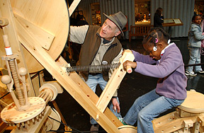 An interactive wood machine created by artist Bernie Ludbell. A little girl straddles the machine and holds handles as if she were riding a bike. A man beside her reaches up, touching a circular structure overhead. (Copyright the Exploratorium, <www.exploratorium.edu> Photo Credit, Amy Snyder.)