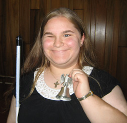 Emily Weidner, smiling, holds a Freedom Bell in one hand and keeps her cane by her side.