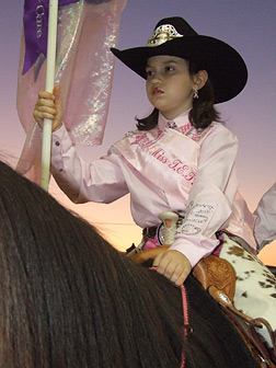 Tacey Raulerson on horseback, holding a flag and dressed in rodeo queen costume. She wears a cowboy hat and a pink pageant sash over a pink western shirt.
