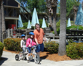 Richard Holloway stands beside a tandem bicycle. Kendra is seated in front and her little brother RJ sits behind her.