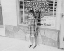 Bonnie as a young child stands outside on the sidewalk in front of her father&rsquo;s insurance business.