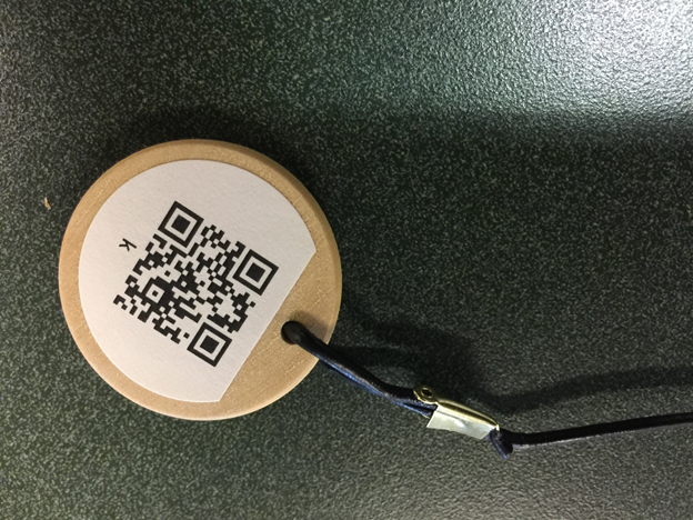 Close-up picture of a wooden coin with a QR code
label attached to it. A leather lanyard is attached to the coin
through a hole drilled in it. The other end of the lanyard is
similarly attached to the 3-D printed point.